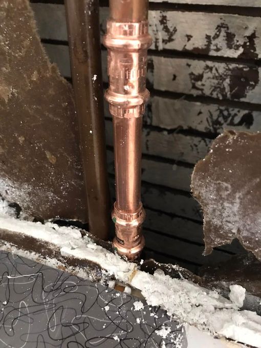 Copper pipes with press fittings run through a hole in a wall, surrounded by crumbling plaster and debris.