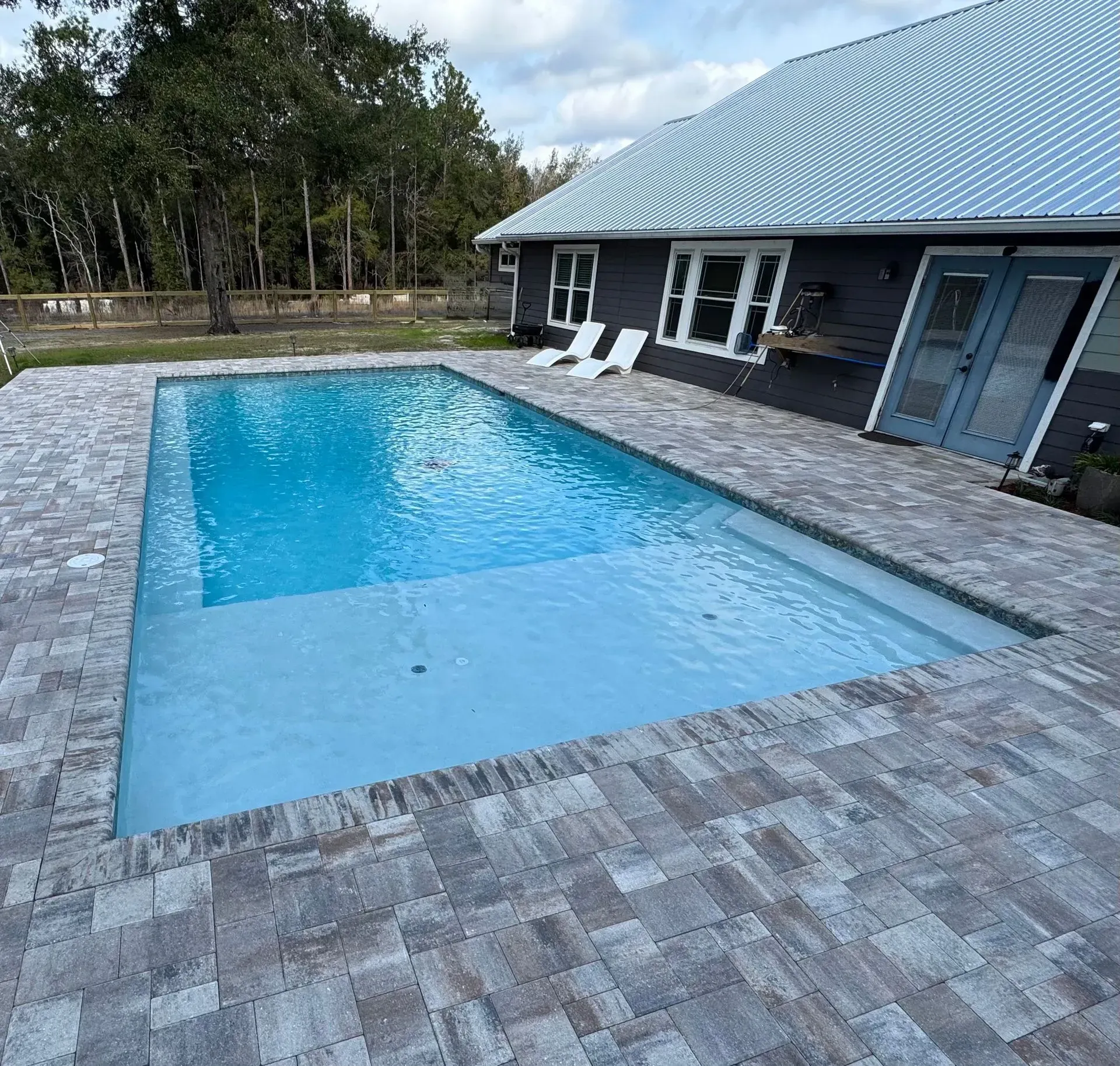 Backyard in-ground pool beside a dark house with a light blue roof and stone paver patio