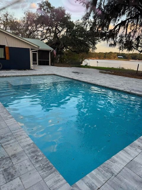 Backyard swimming pool with blue water beside a house, trees, and a view of the water at sunset