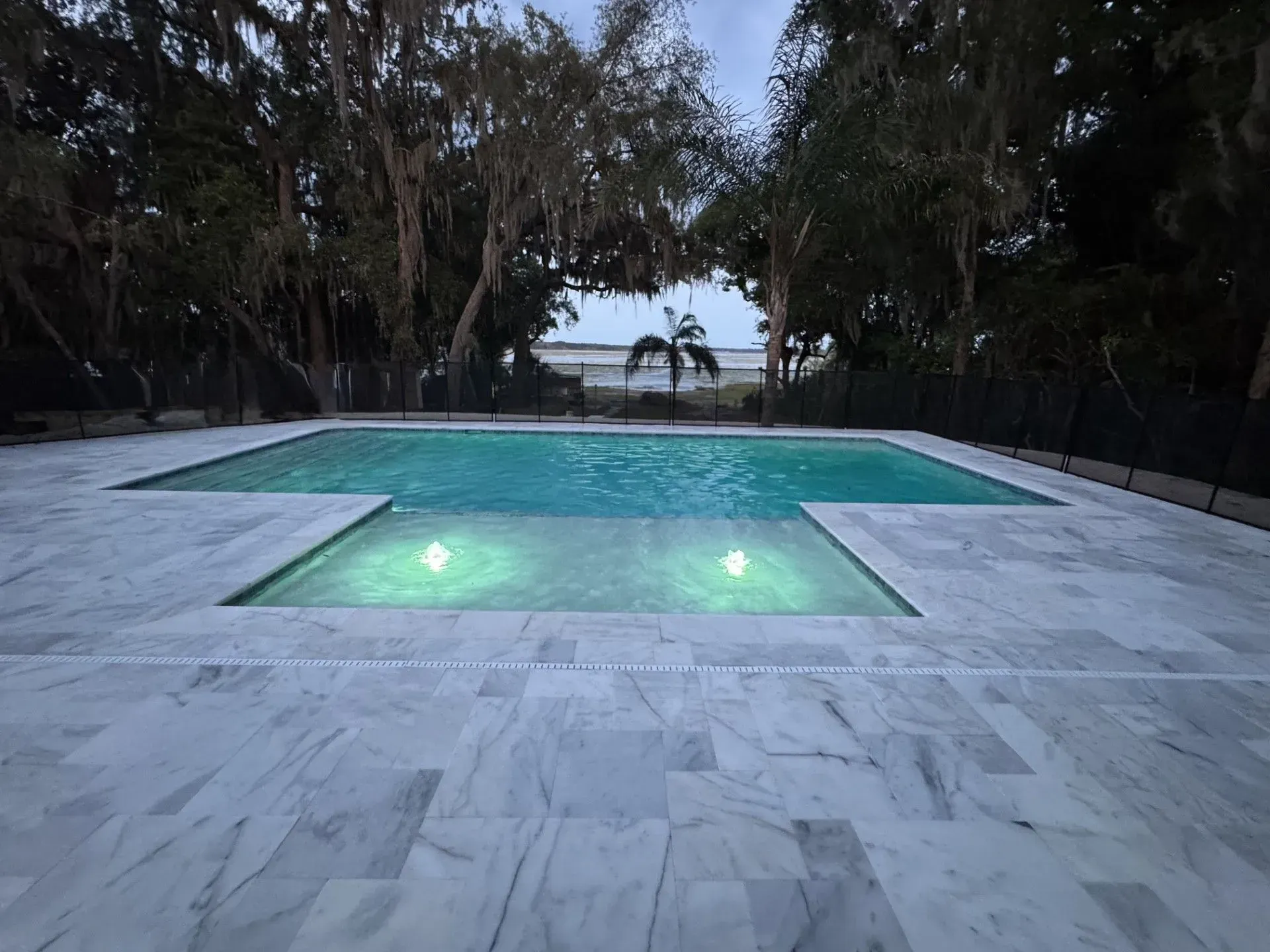 Empty backyard pool with glowing underwater lights at dusk, framed by palm trees and ocean view