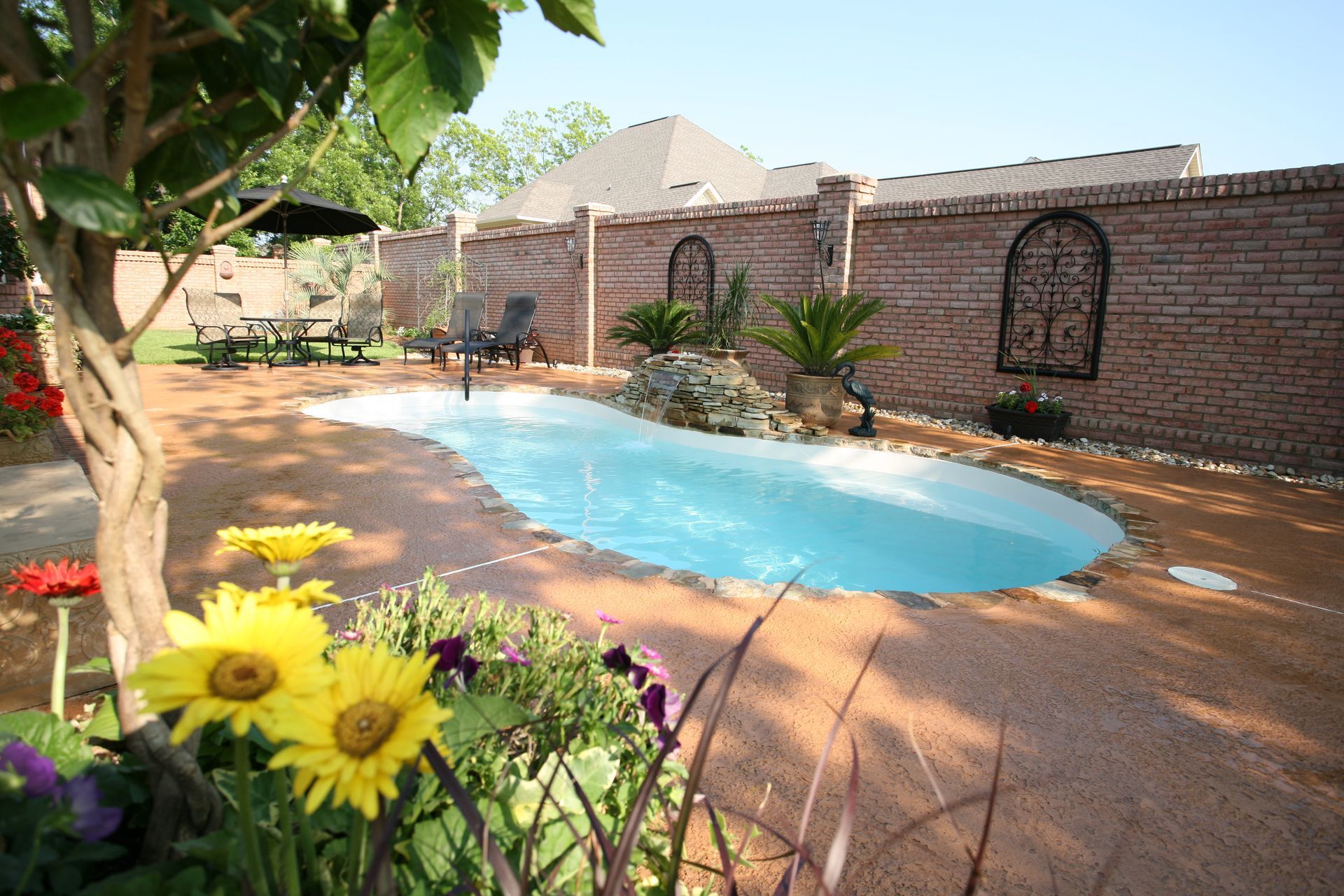 Backyard swimming pool beside a brick wall, with yellow flowers in the foreground and a tree shading the patio.