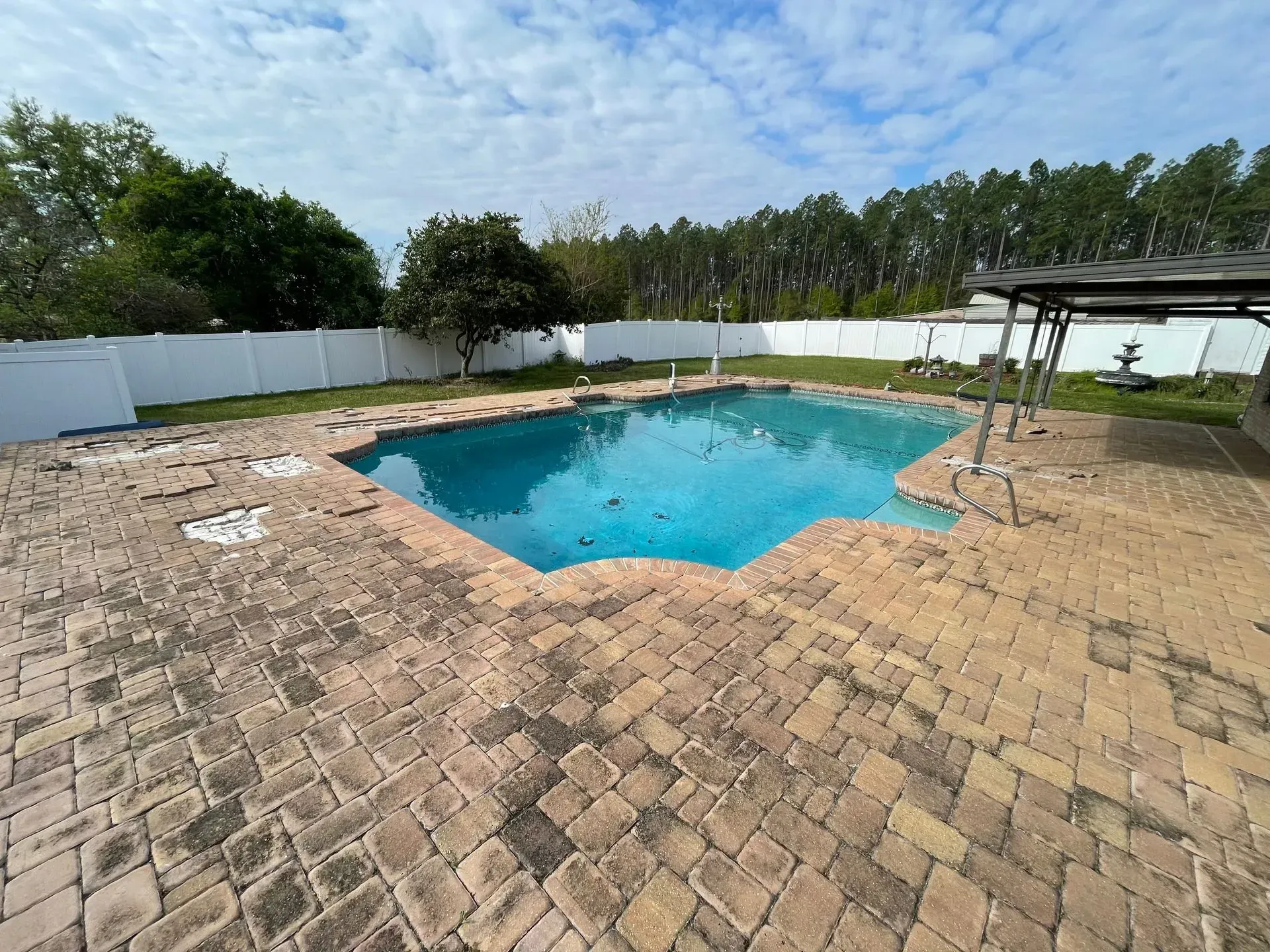 Backyard pool with blue water, brick patio, white fence, and trees under a partly cloudy sky