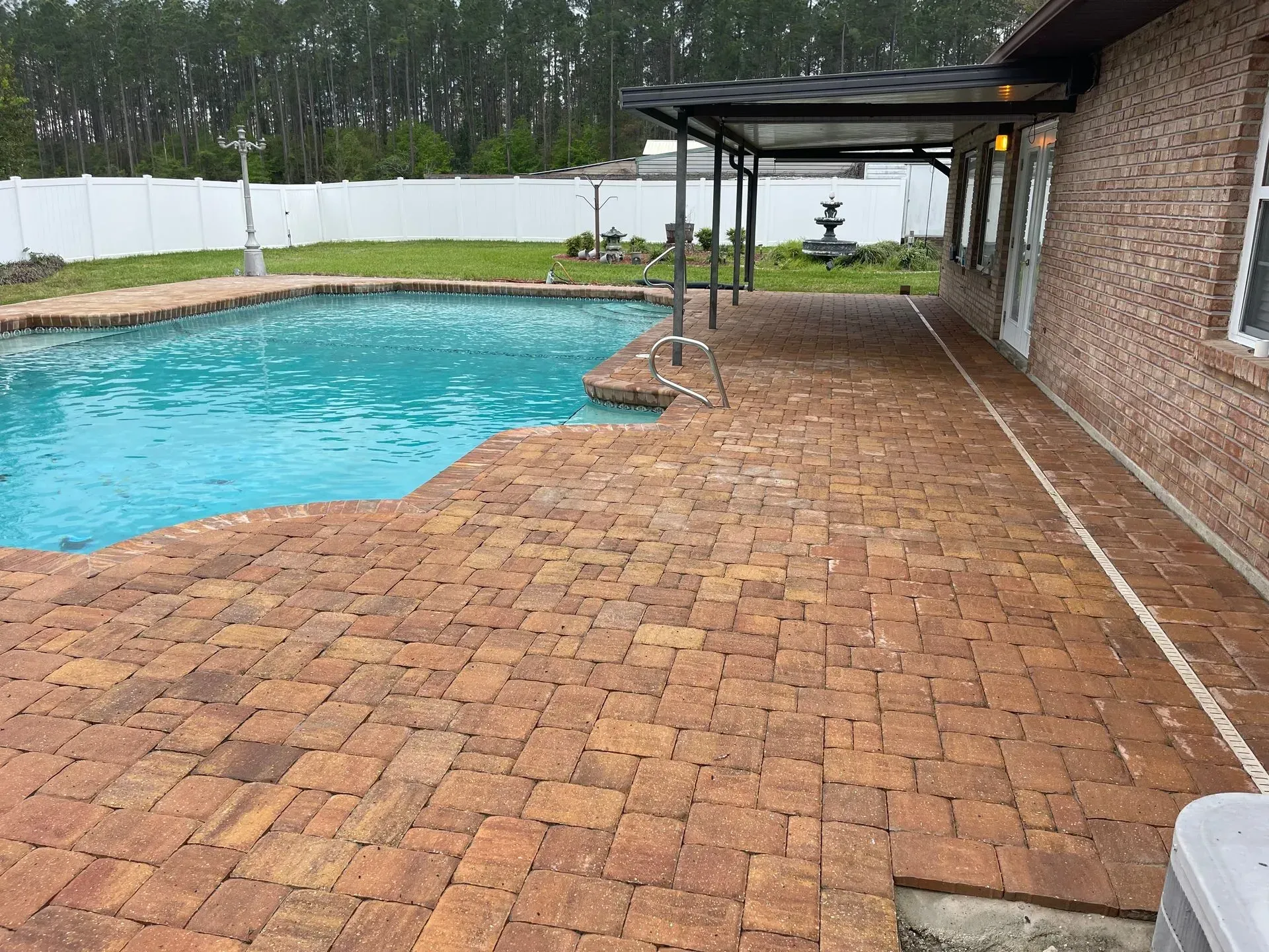 Brick pool deck beside a turquoise swimming pool, with a covered patio and white fence in the background