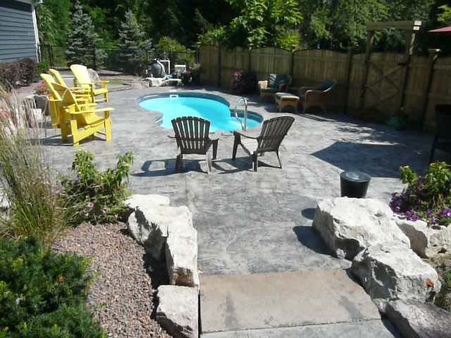 Backyard patio with a pool, chairs, stone path, and landscaped garden under bright sunlight