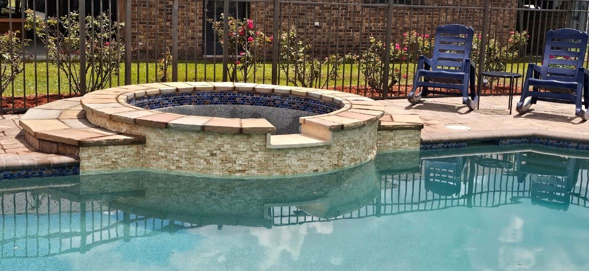 Pool with built-in hot tub. Two blue lounge chairs sit nearby, with a wrought iron fence and bushes in the background.