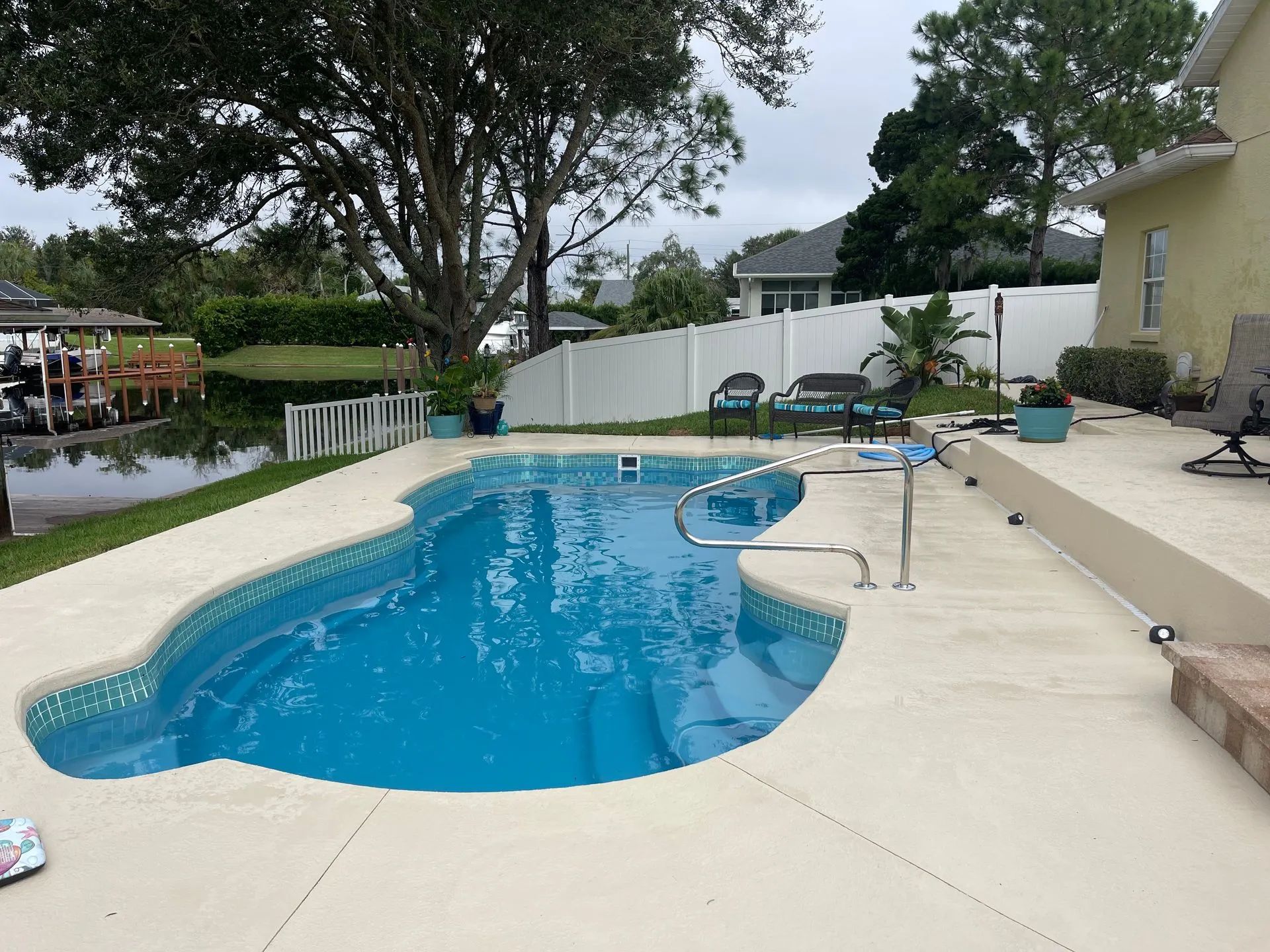 Backyard swimming pool with diving board, lounge chairs, and trees along a fenced patio