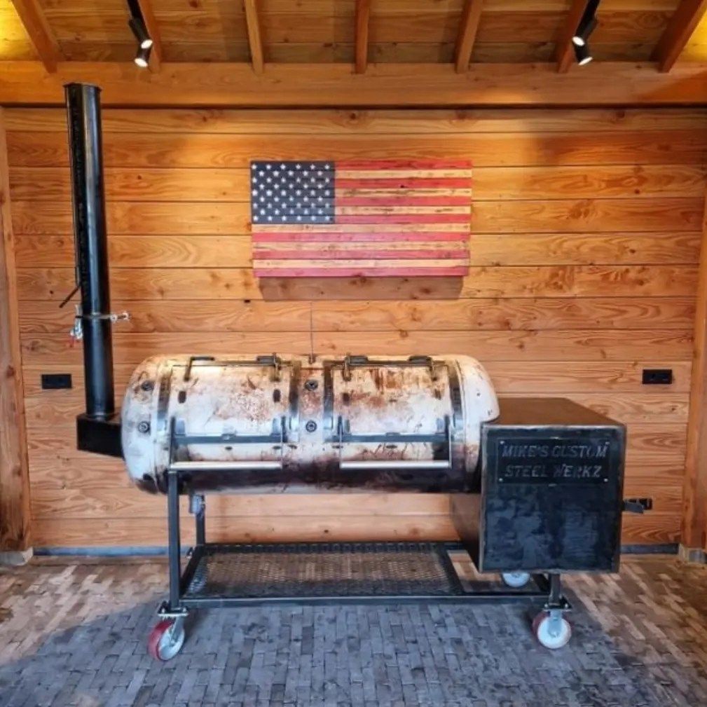 A bbq grill is sitting in front of a wooden wall with an american flag above it.