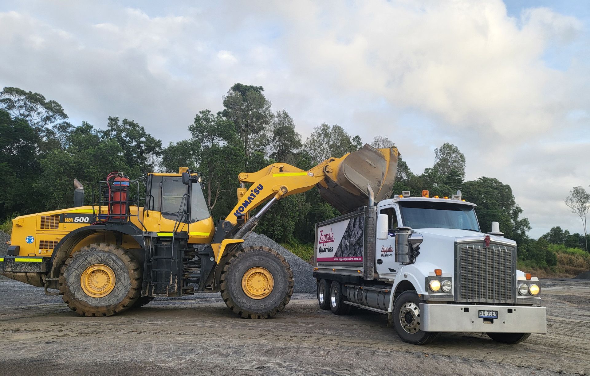 Yellow loader dumping gravel into a white dump truck, outdoors. Cloudy sky, trees in the background  — Zappala Quarries in Gordonvale, QLD