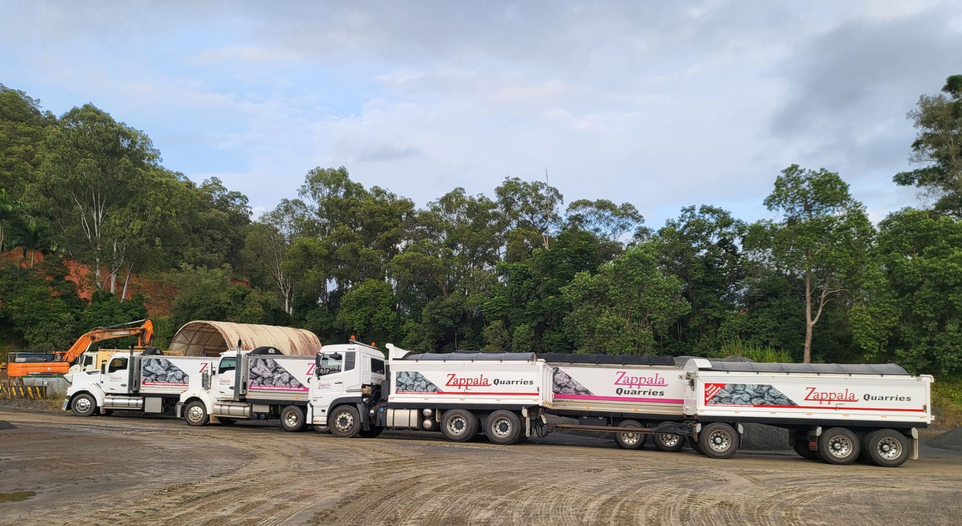 A line of white dump trucks with gravel in them, parked in a dirt lot by a wooded hill — Zappala Quarries in Gordonvale, QLD