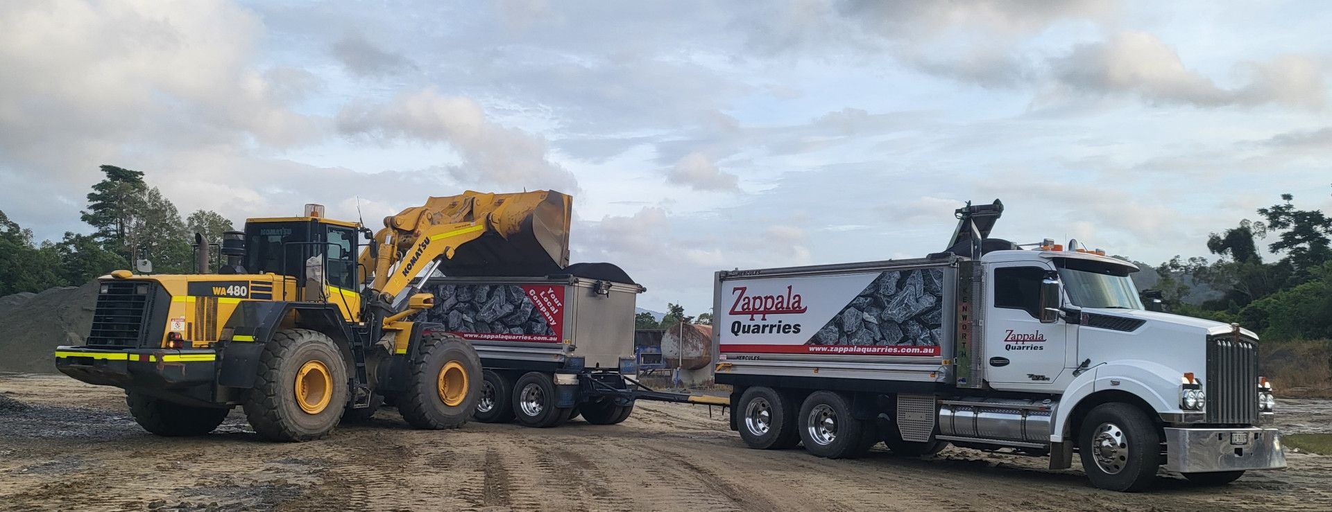 A yellow loader fills a white dump truck with gravel at a construction site under a cloudy sky — Zappala Quarries in Gordonvale, QLD