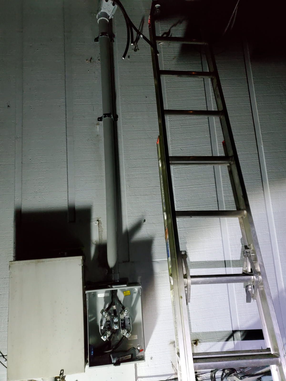 Ladder leaning against a wall with electrical equipment; conduit and cables are attached to the wall.