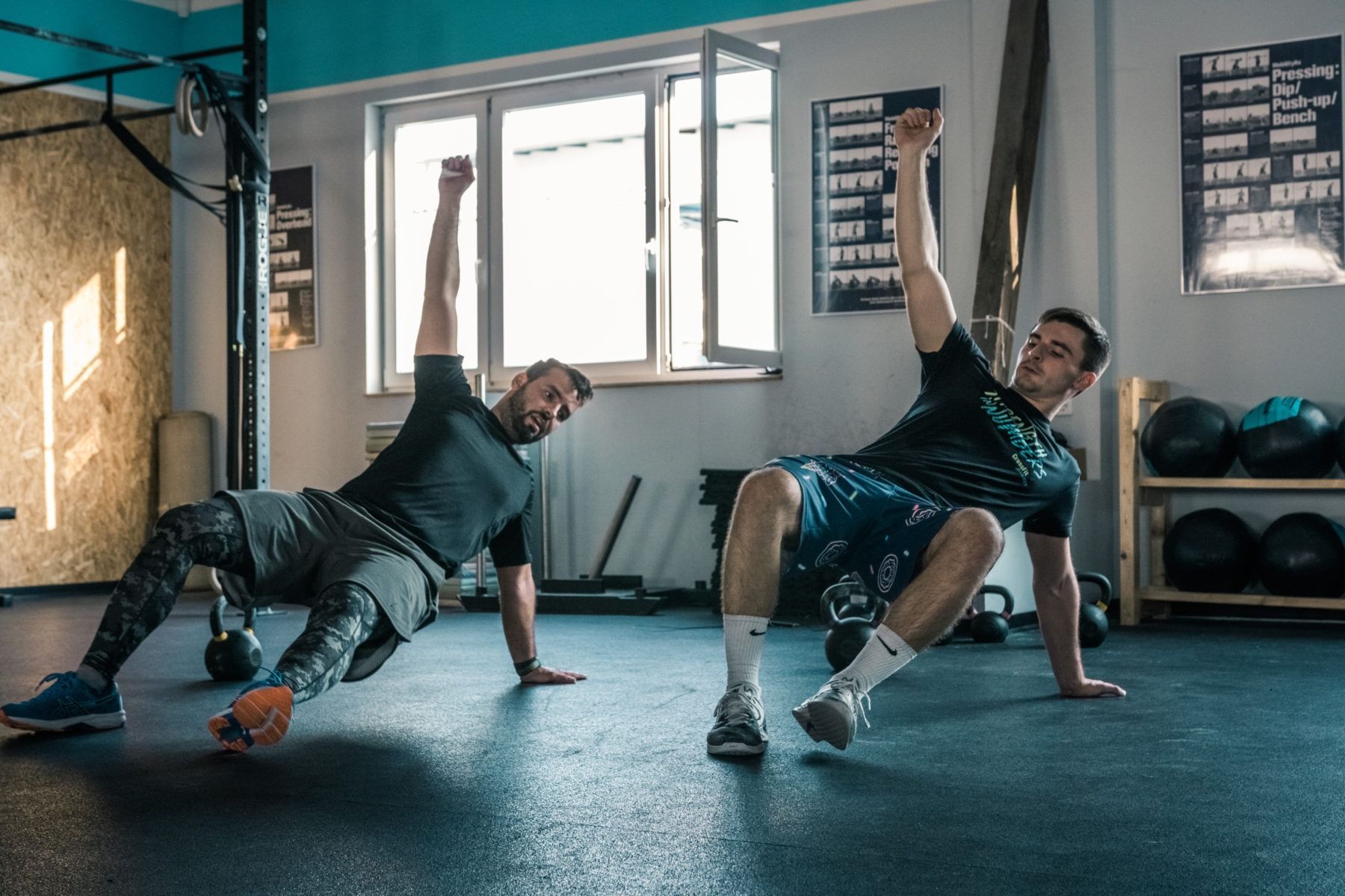 Two individuals in a gym performing a side plank exercise with one arm raised toward the ceiling.