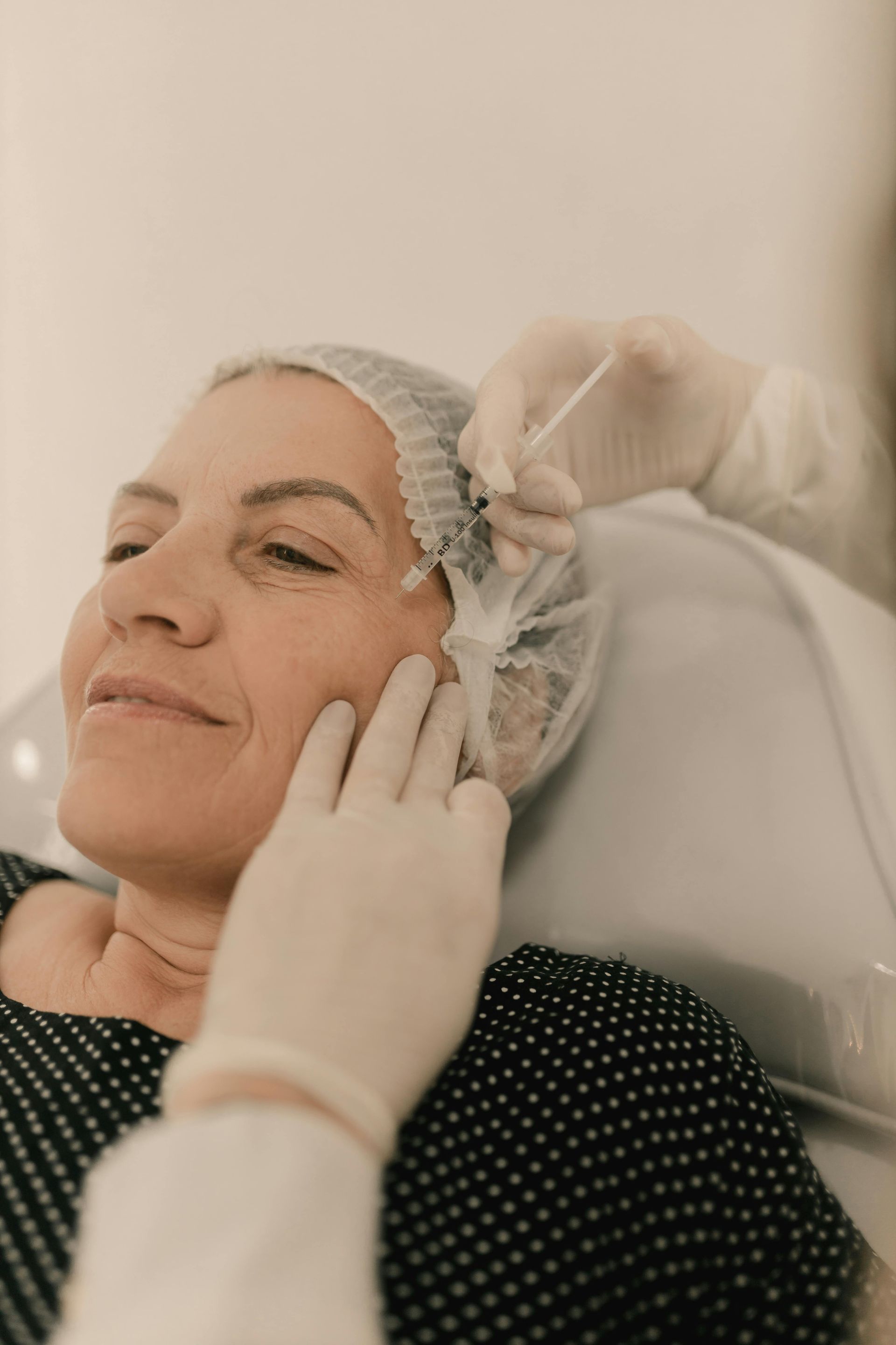 A gloved clinician applies a facial treatment to a relaxed patient wearing a hair cap.