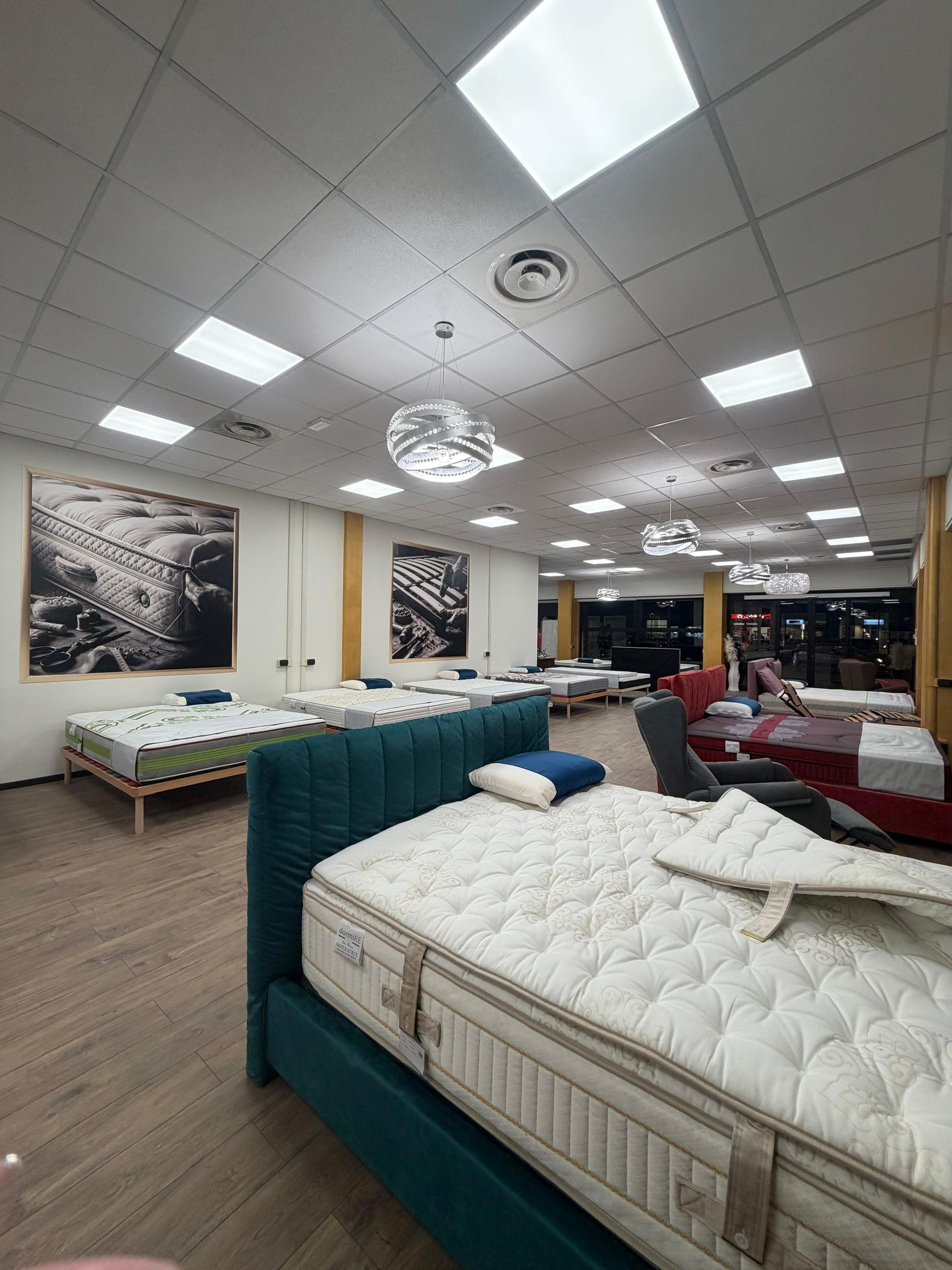 Interno di un negozio di materassi con numerosi letti esposti; illuminazione dall'alto; opere d'arte alle pareti.