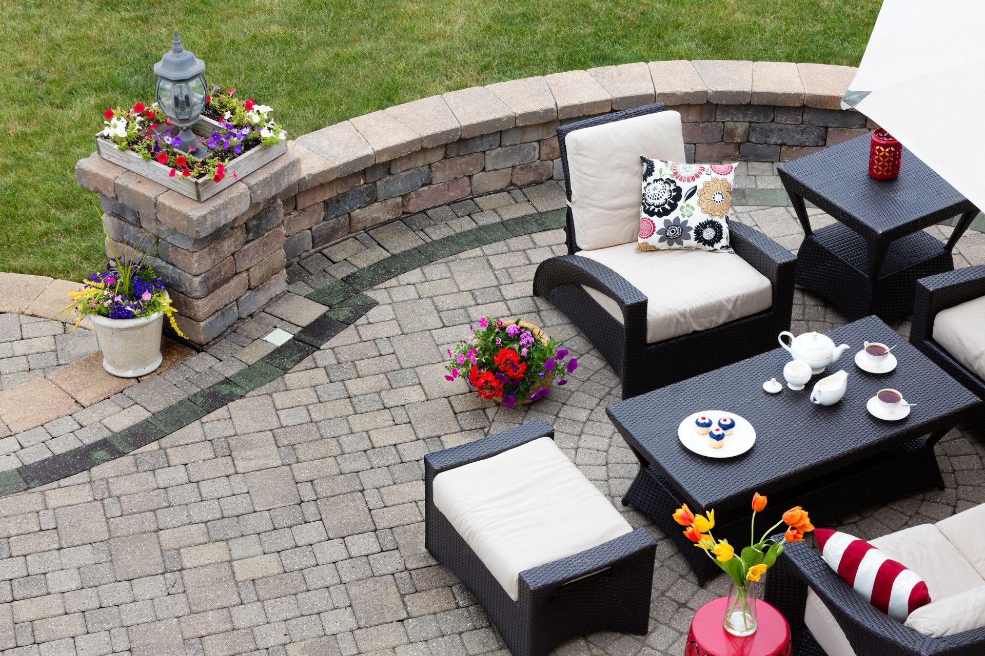 Patio with outdoor furniture, flowers, and brick wall. Black wicker chairs with cream cushions, stone patio.