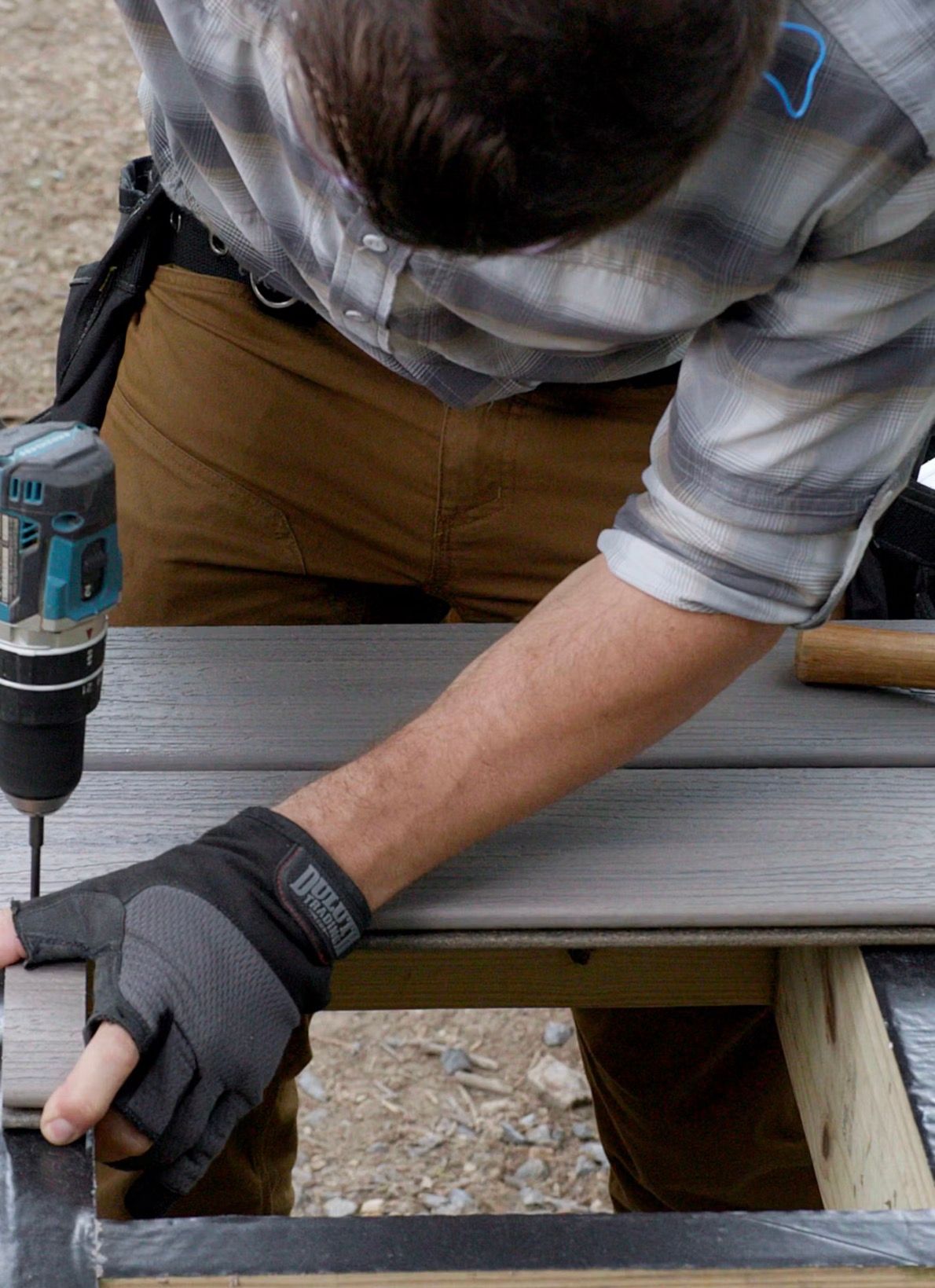 Person using a drill to screw a board onto a deck frame, wearing gloves.