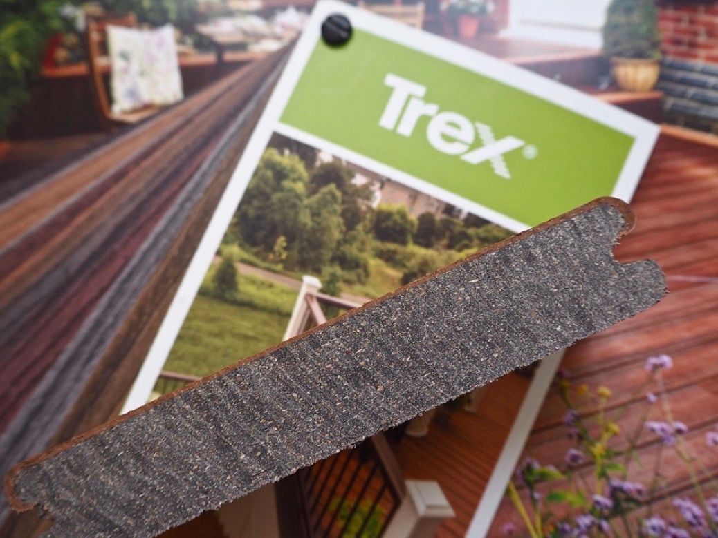 Cross-section of Trex composite decking board; dark brown with a textured surface, placed over a brochure.