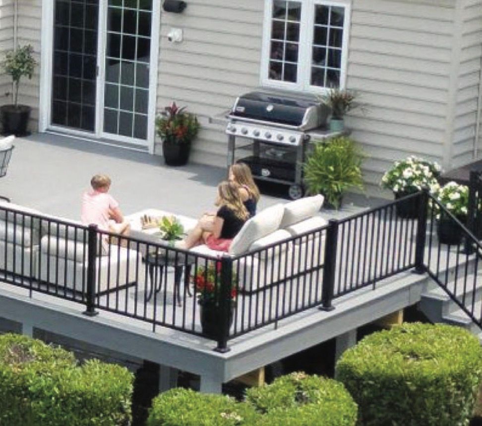 People on a gray deck with black railing, sitting on white outdoor furniture. A grill, potted plants, and the house are in view.