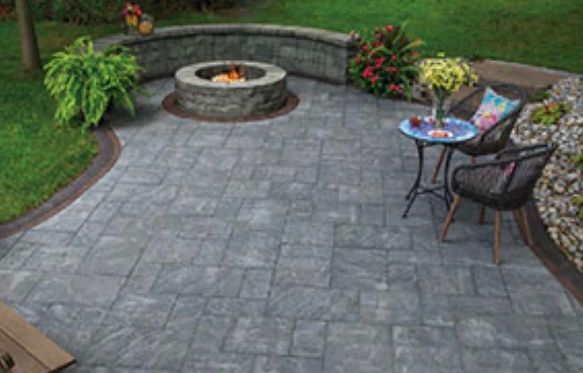 Patio with gray stone pavers, fire pit, table and chairs, surrounded by greenery.