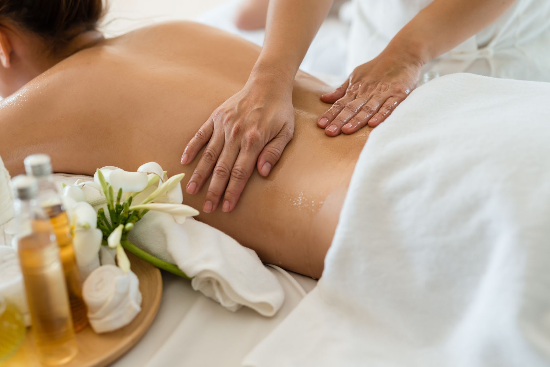 A massage therapist performs a back massage on a client lying on a white massage table with flowers and oils nearby.
