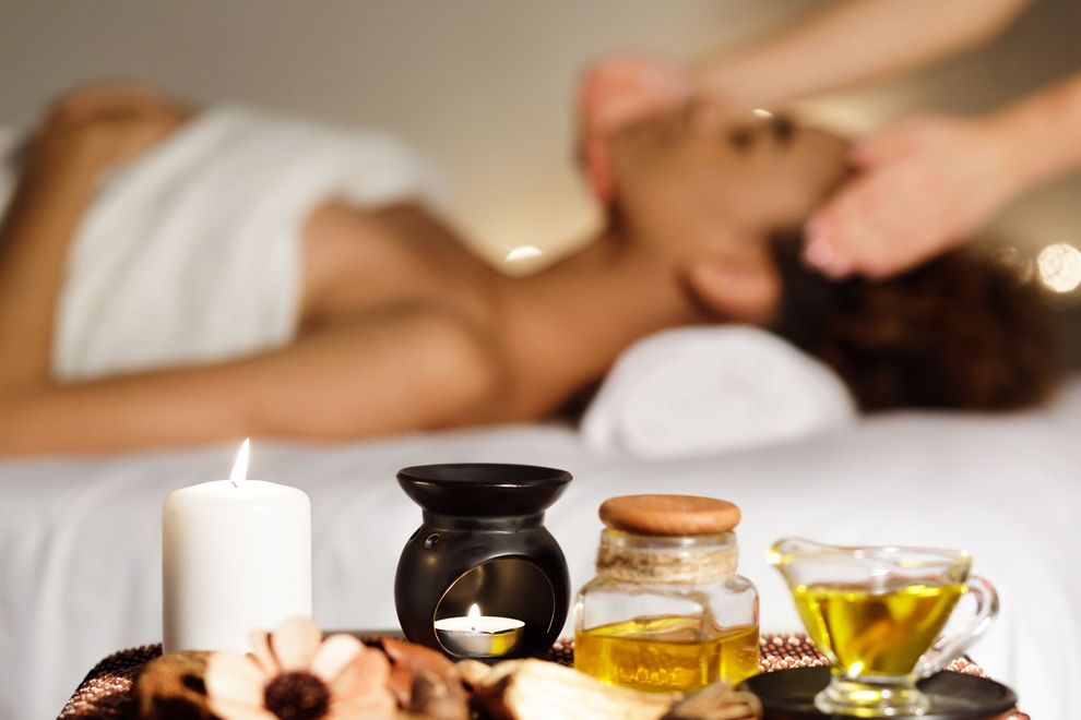 A reed diffuser, two lit candles, stacked grey stones, and white plumeria flowers on a wooden table in a massage setting. A reed diffuser, two lit candles, stacked grey stones, and white plumeria flowers on a wooden table in a massage setting.
