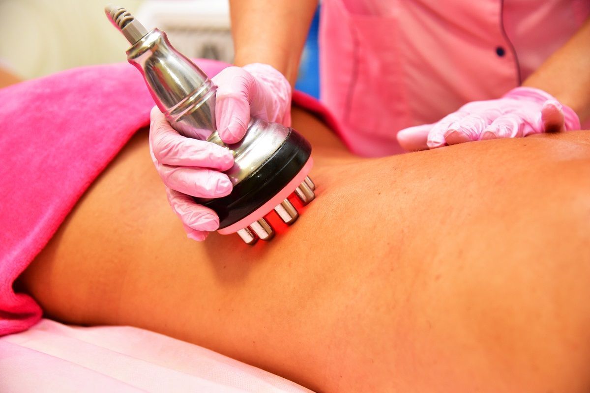 A practitioner in pink gloves uses a handheld cosmetic device on a person's lower back during a skin treatment.