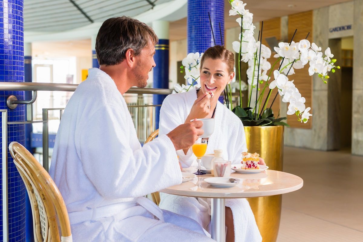 Two people in white bathrobes sit at a small round table, drinking juice and eating, in a modern, plant-filled spa area.