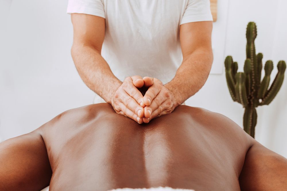 A massage therapist performing sports massage deep tissue work on a person's upper back near a cactus in a bright, modern room.