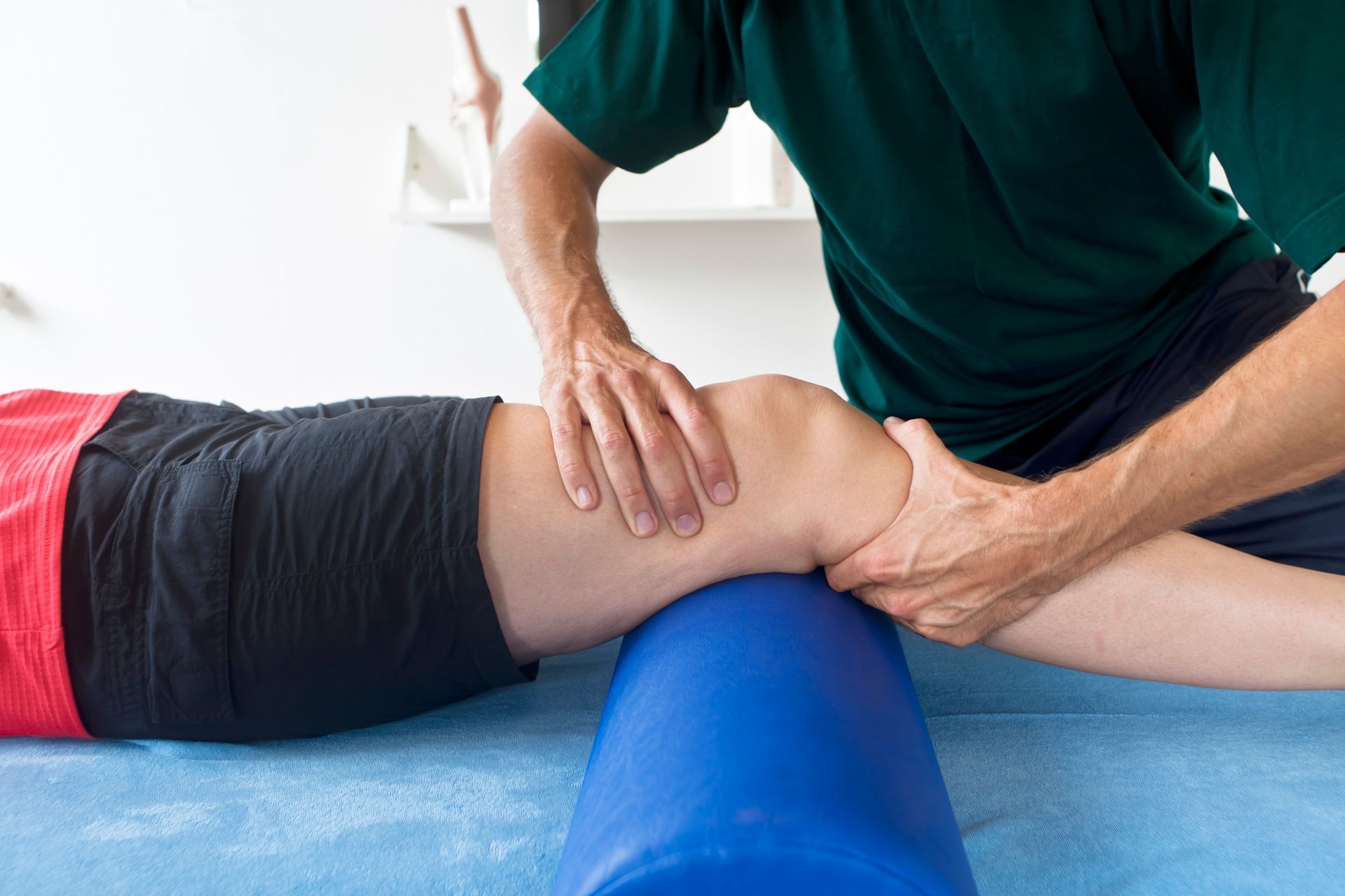 A physical therapist performs knee joint mobilization on a patient's leg, supported by a blue foam roller on a blue table.