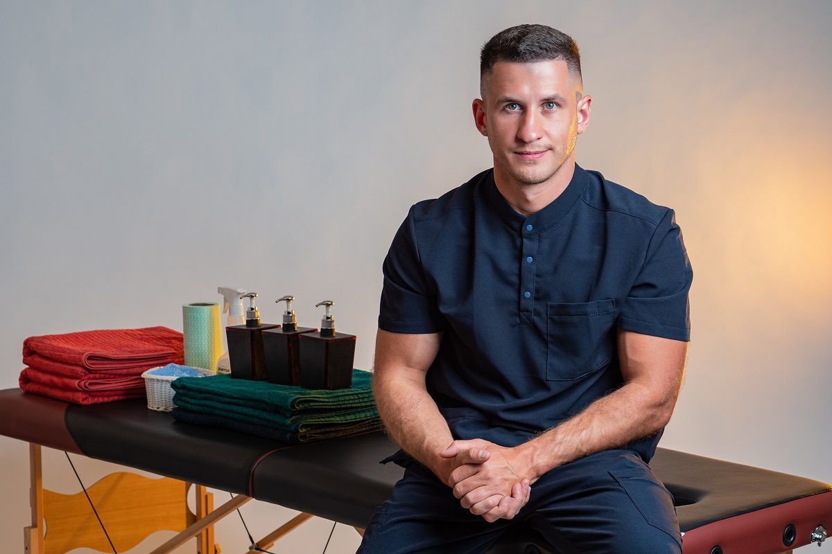 A person in dark blue scrubs sits on a massage table next to folded towels and pump bottles in a neutral-toned room.