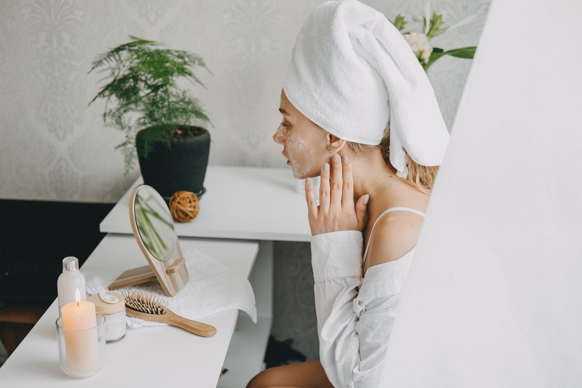 A person with a white towel wrapped around their head touching their face in front of a small mirror on a desk.