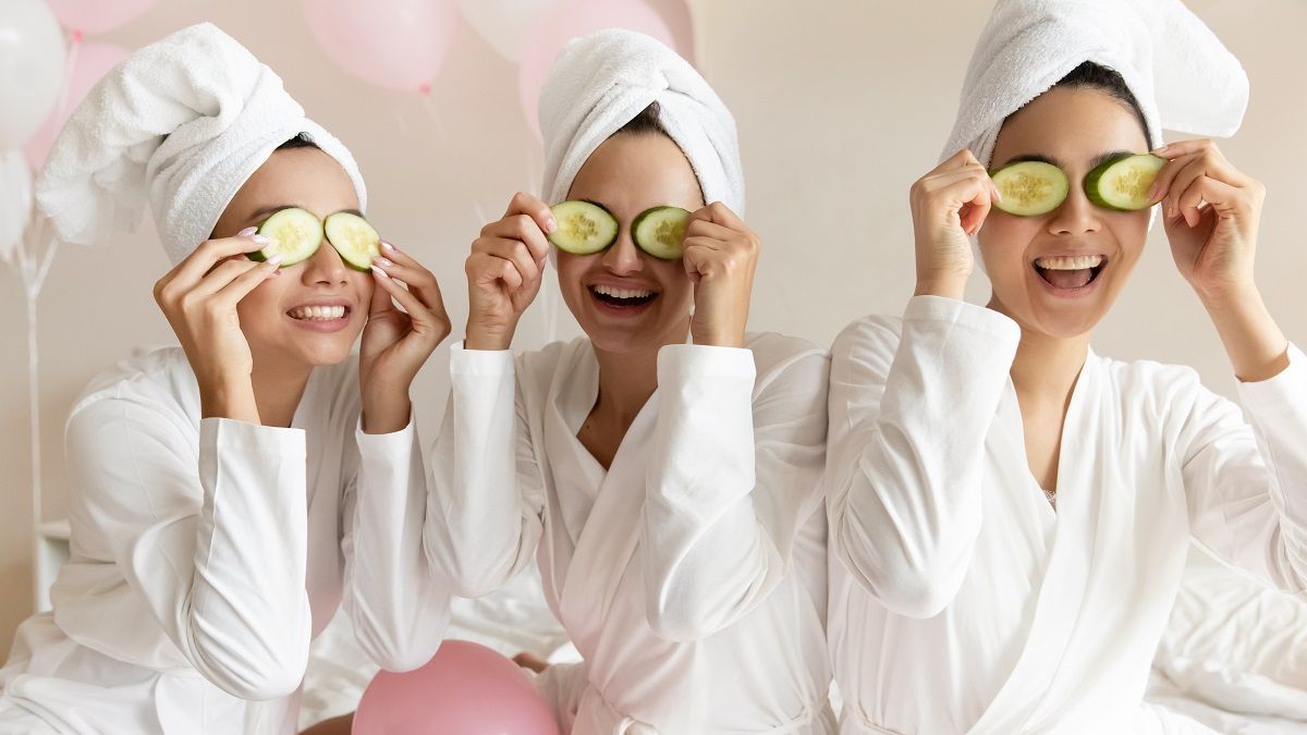 Three people in white robes and hair wraps hold cucumber slices over their eyes, smiling in a room with pink balloons.