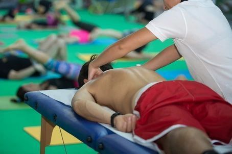 A person in a white shirt performs a neck massage on an individual lying face down on a massage table at a gym.