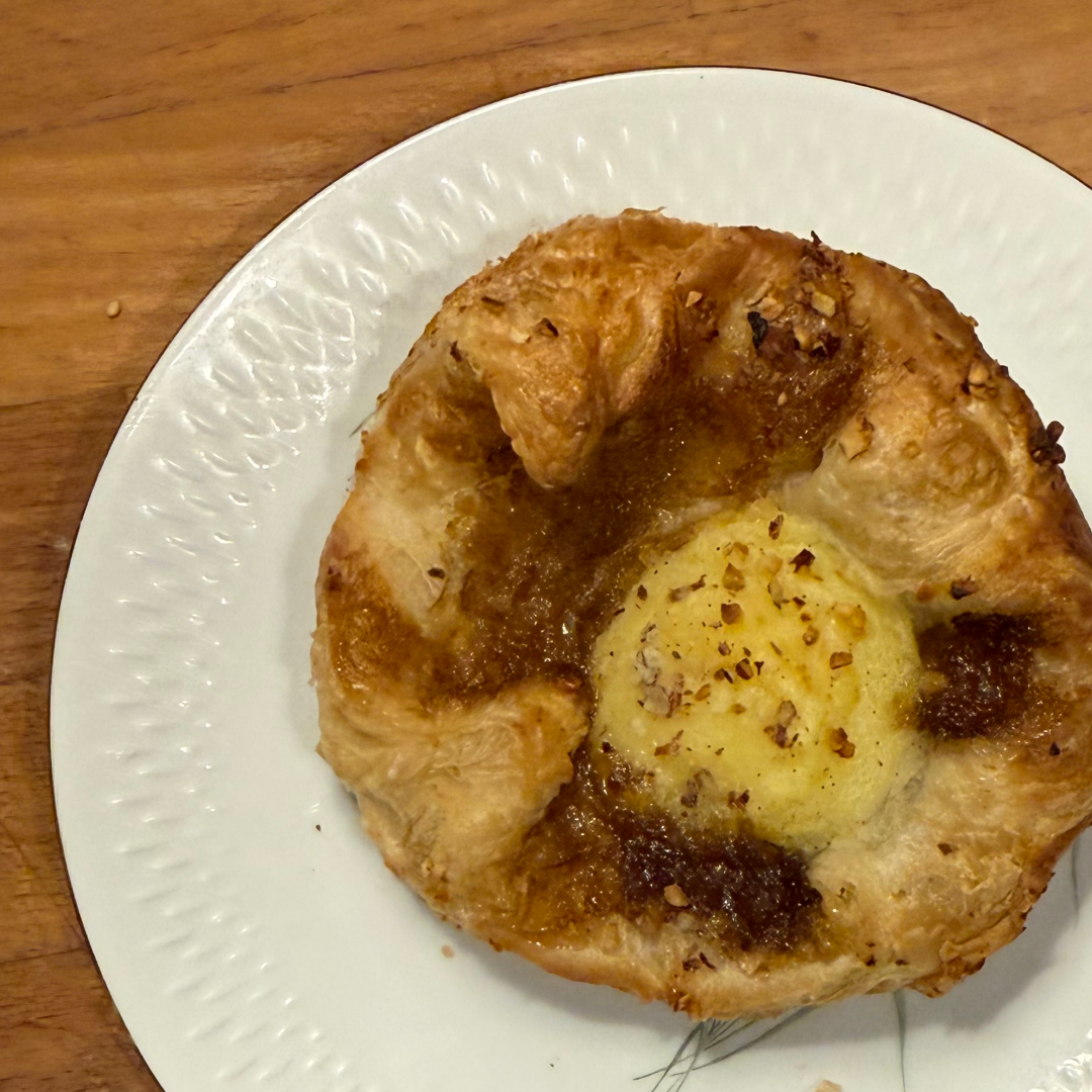 En rund wienerbrød med en gul midte og brunede kanter på en hvid tallerken på et træbord.