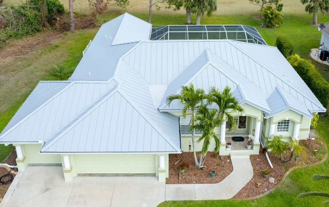 Aerial view of a light green house with a light gray metal roof, a screened pool, and a landscaped yard.