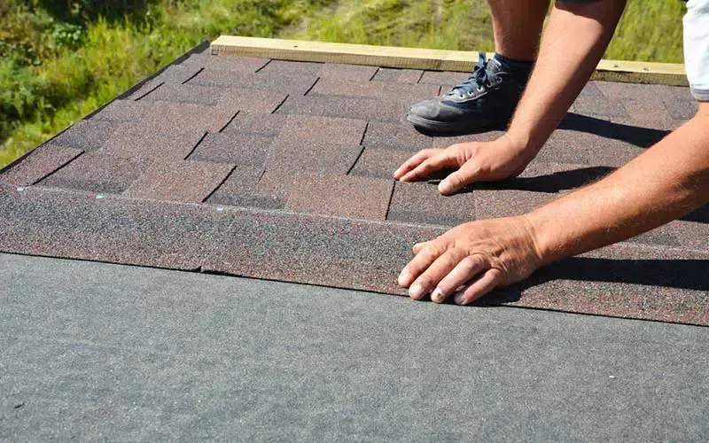 Person installing brown shingles on a roof, hands visible.