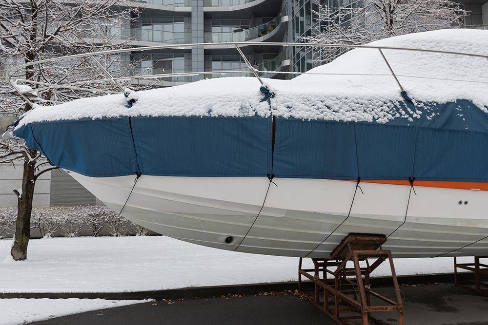 Boat covered in blue tarp, coated with snow, on stands in snowy environment, near a building.