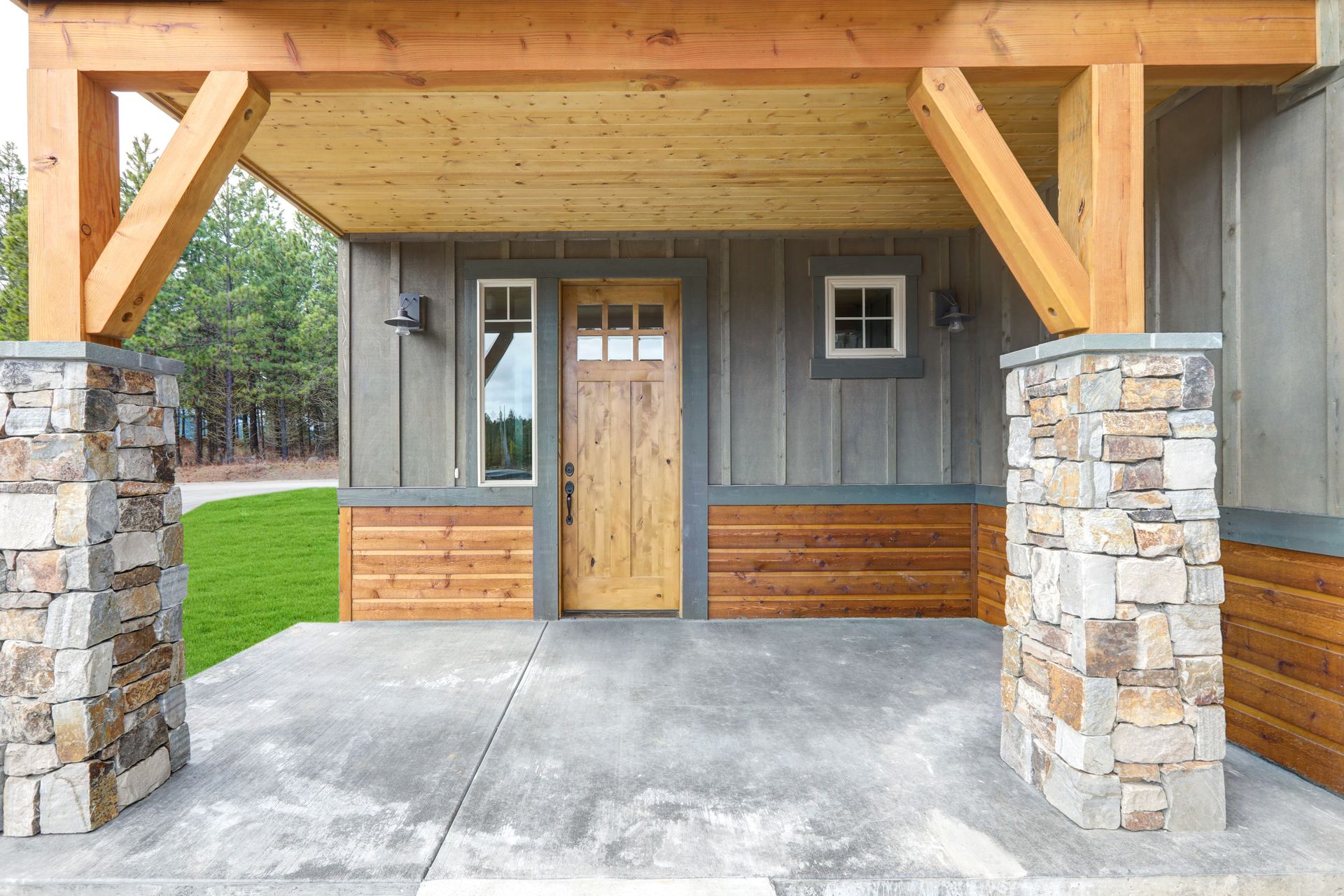 Porch with stone columns and wooden beams. Brown door, gray siding, and concrete floor.