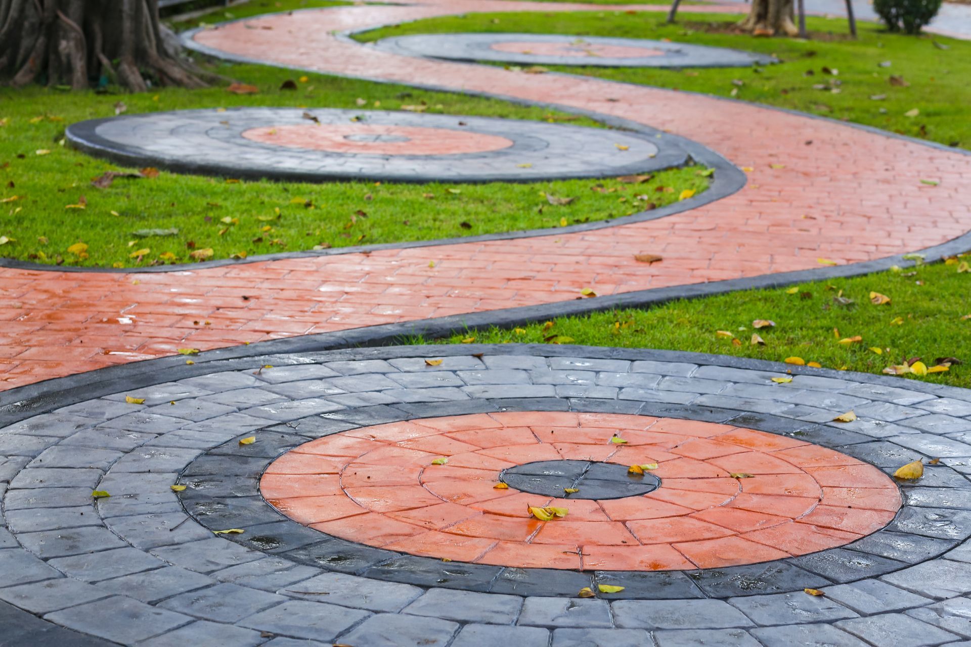 Winding brick path with circular insets of red, black, and gray on grassy ground in a park.