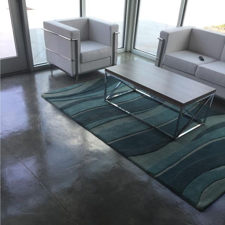 Modern living room with white furniture, a blue rug, and a silver coffee table, beside large windows.