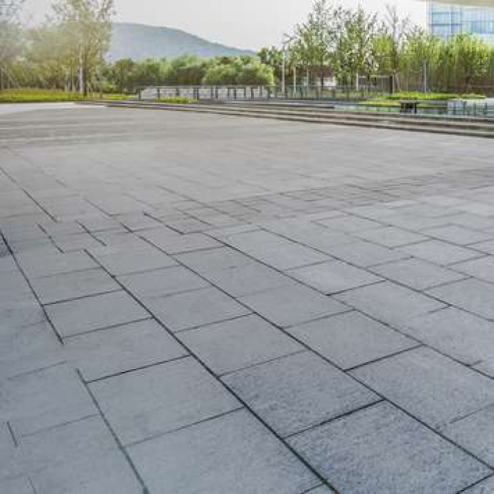 A gray paved plaza with rectangular stones, trees, and a building in the background.