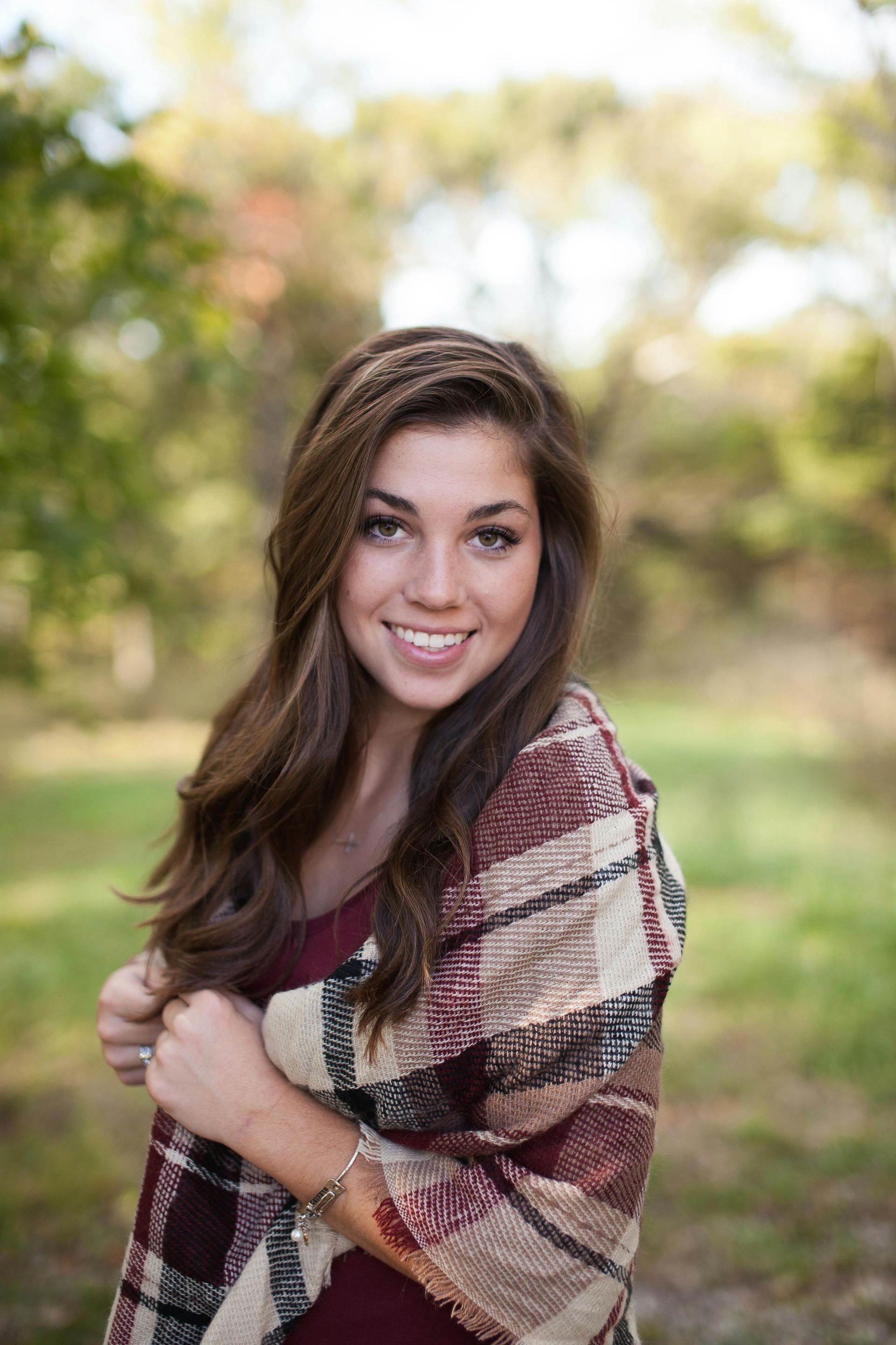 Young woman smiling, wrapped in a plaid shawl, standing outdoors with blurred foliage background.