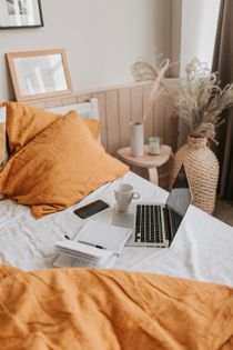 Bed with laptop, coffee, and notepad, bathed in warm, natural light. Orange pillows and blanket; neutral-toned decor.