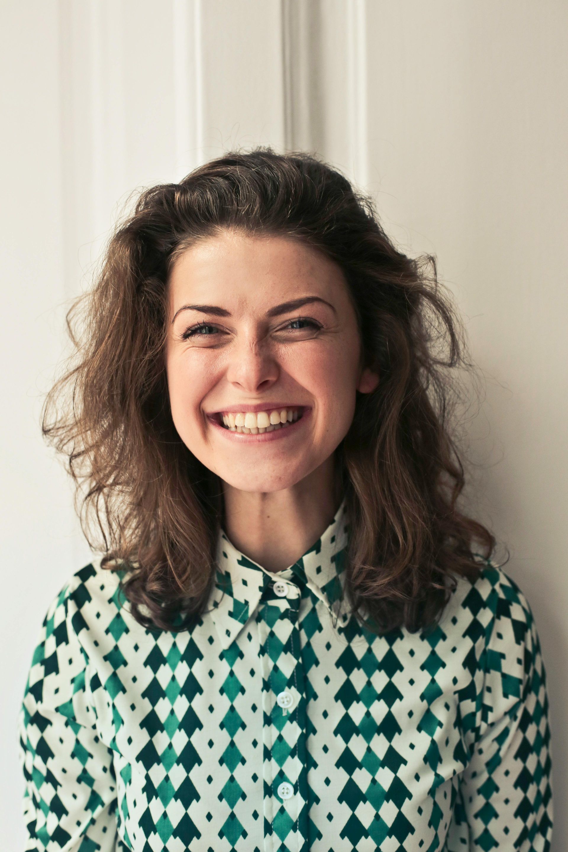Woman with curly brown hair, wearing a patterned shirt, smiles broadly in front of a white door.