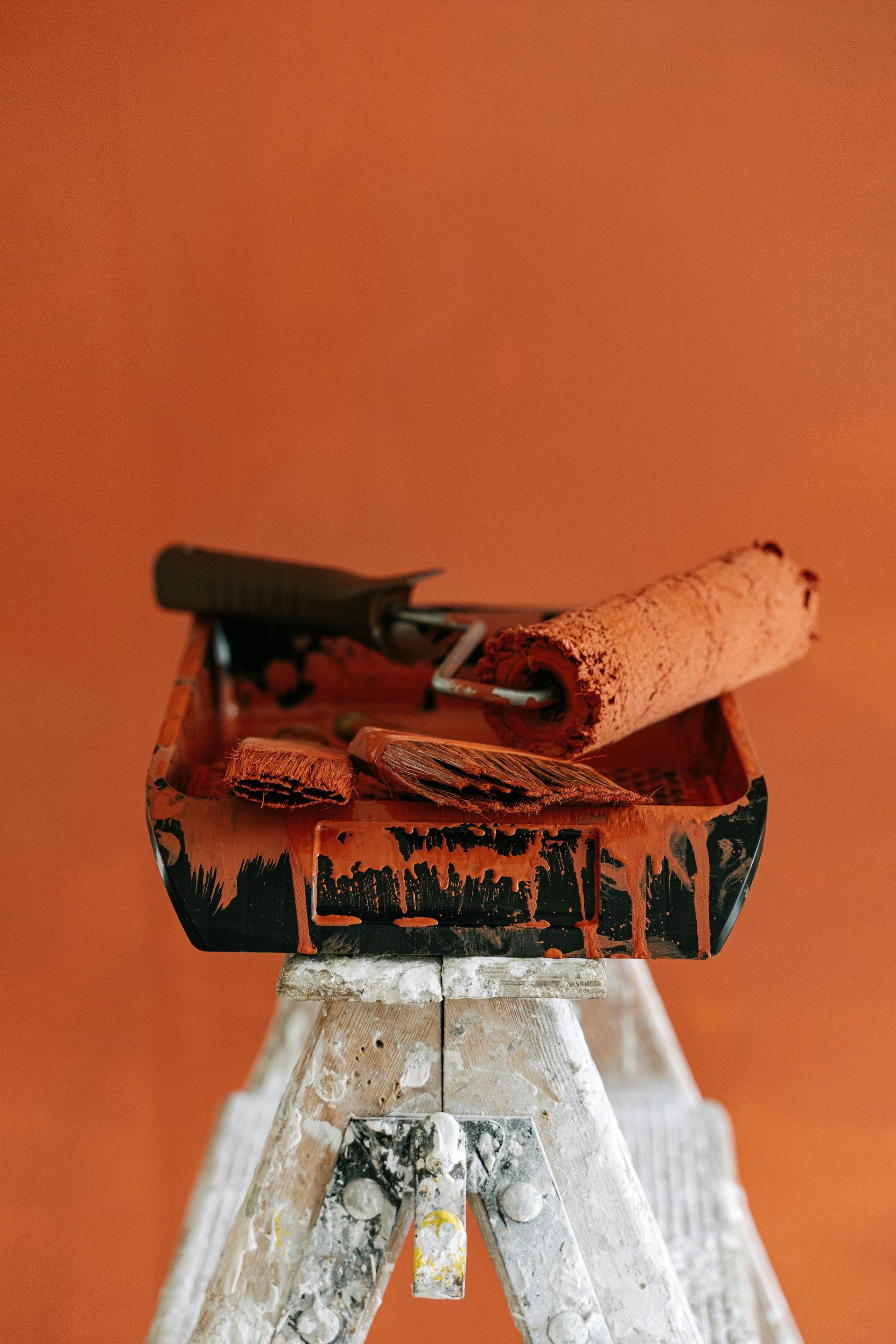 Paint tray with roller and brush on a small ladder, all covered in reddish-orange paint.