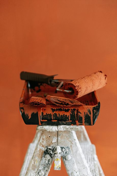Paint tray with roller and brush on a small ladder, all covered in reddish-orange paint.