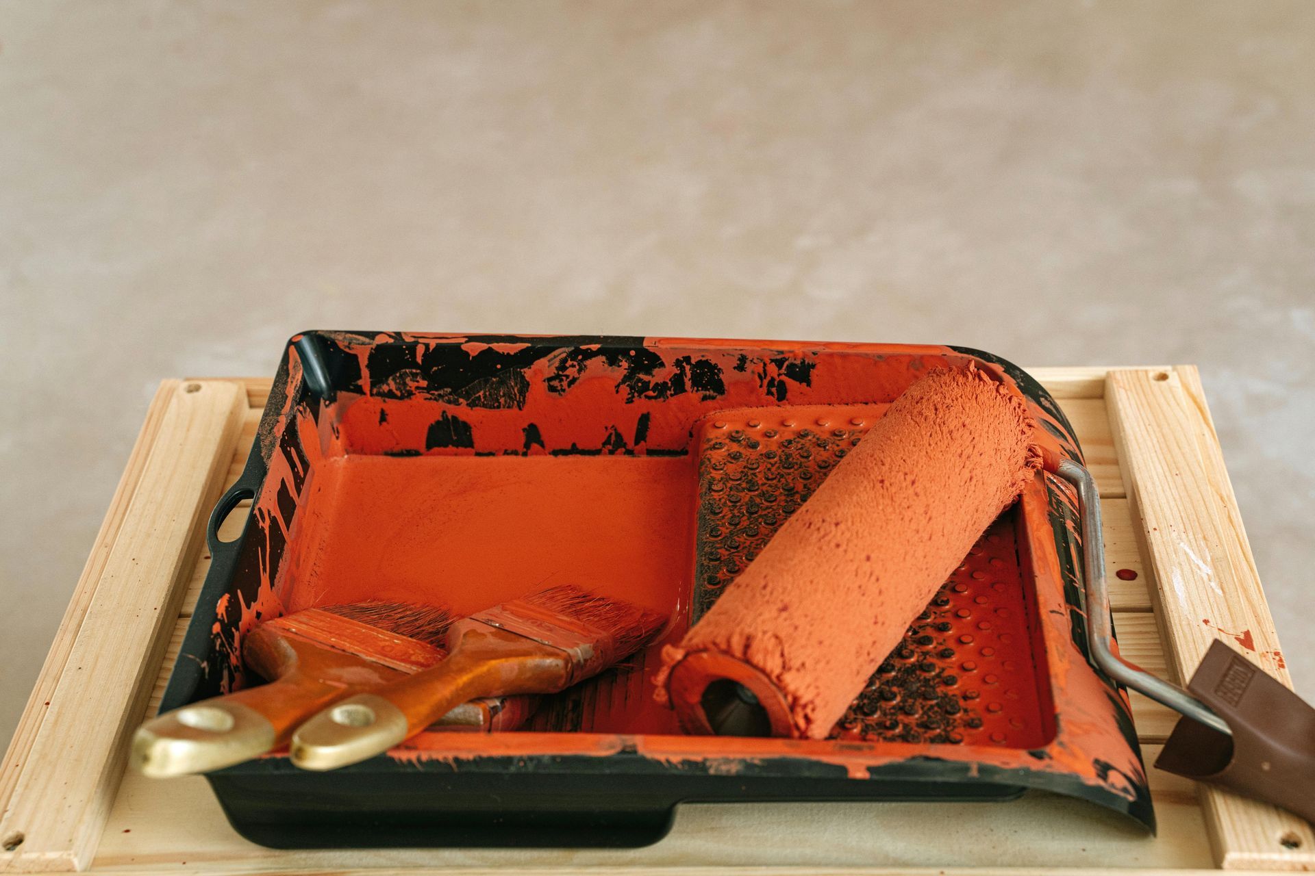 Orange paint supplies in tray on wooden surface: brushes, roller, and scraper.