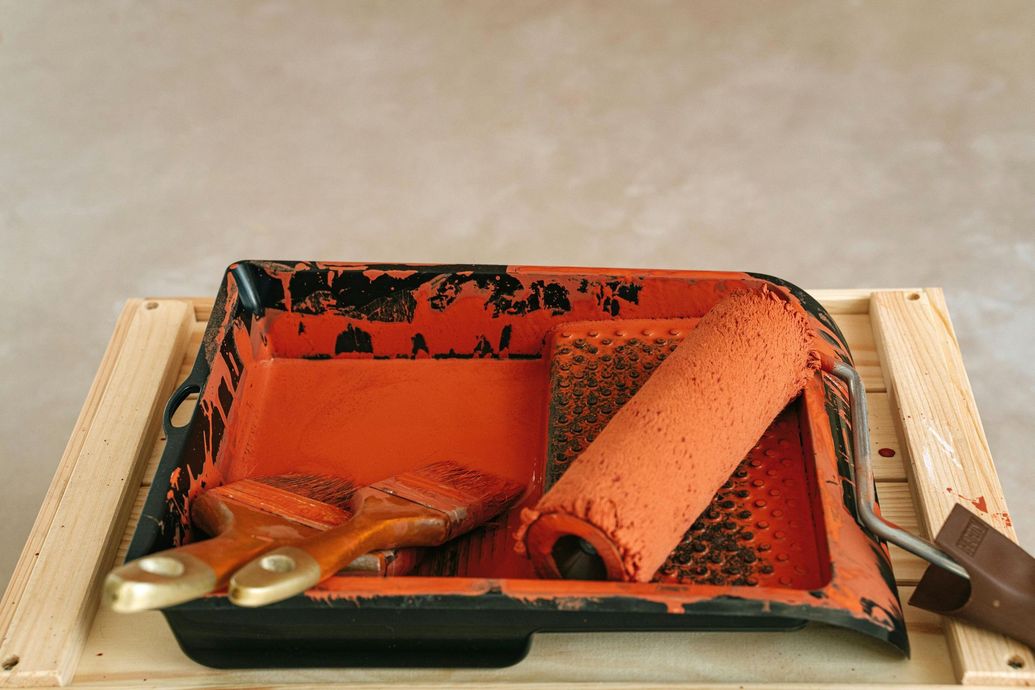 Orange paint supplies in tray on wooden surface: brushes, roller, and scraper.
