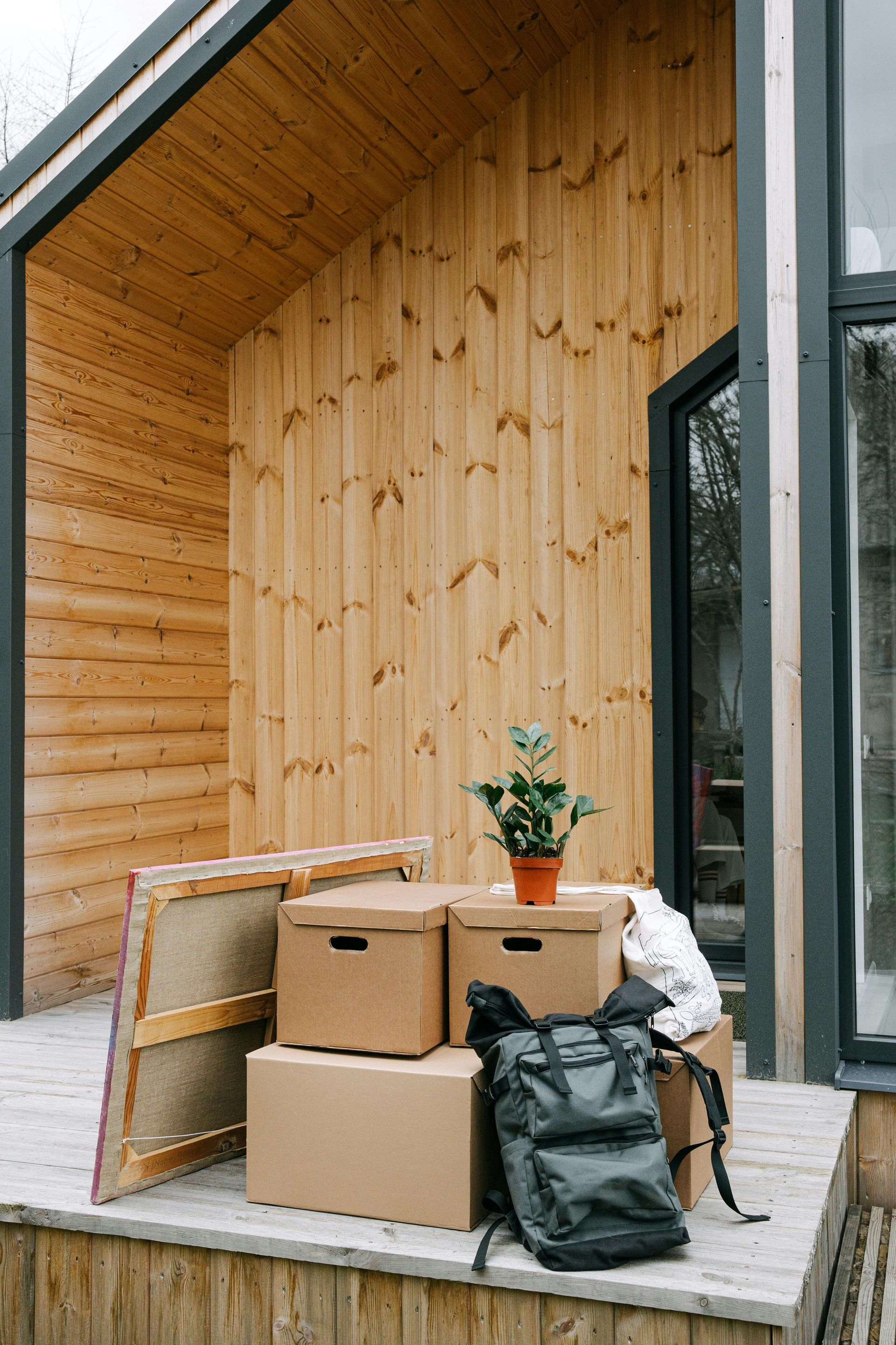 Cardboard boxes, backpack, and plant on a porch in front of a wood-paneled building; moving day.
