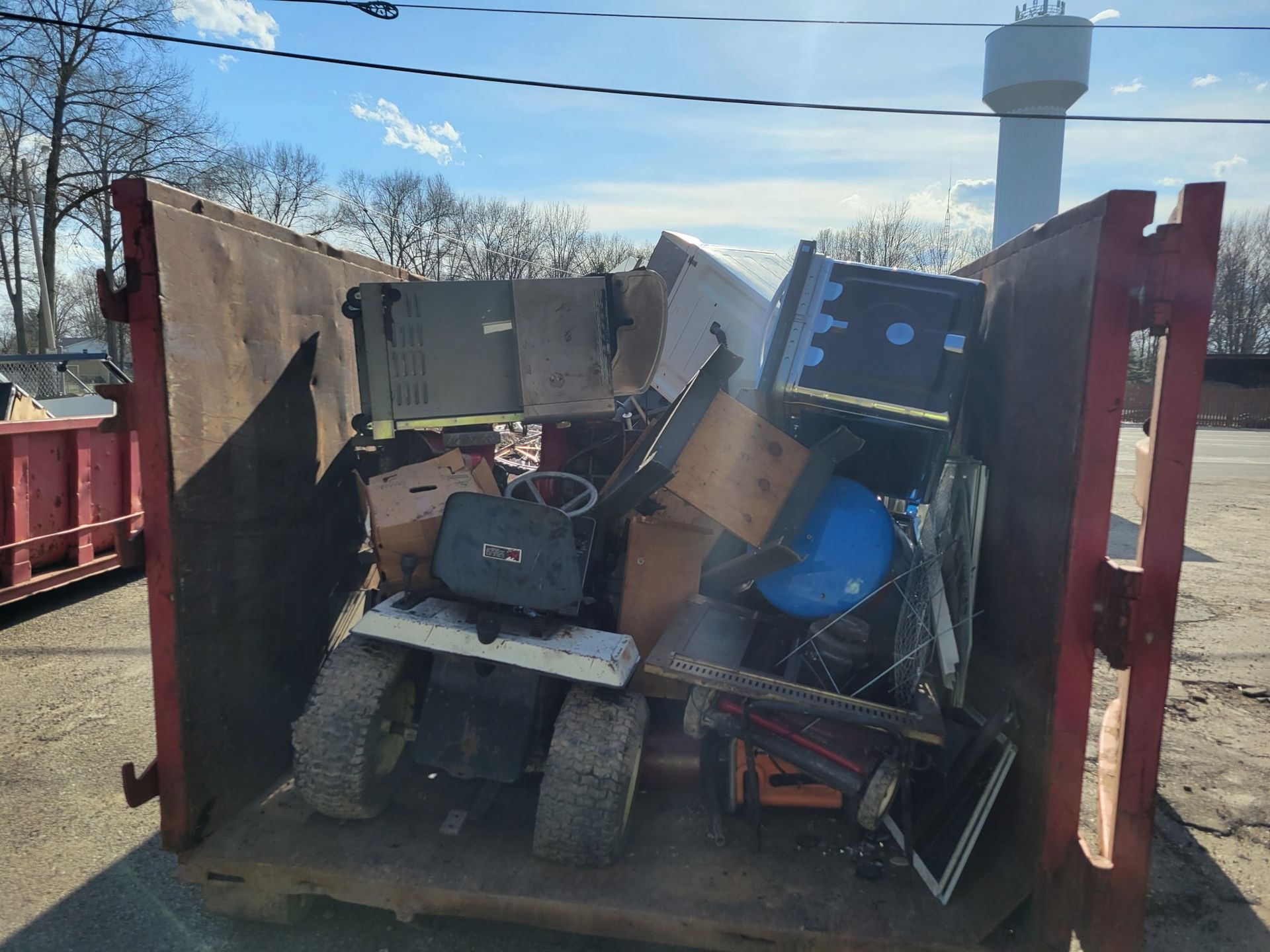 A red dumpster filled with discarded items including a lawnmower, computer, and wood.